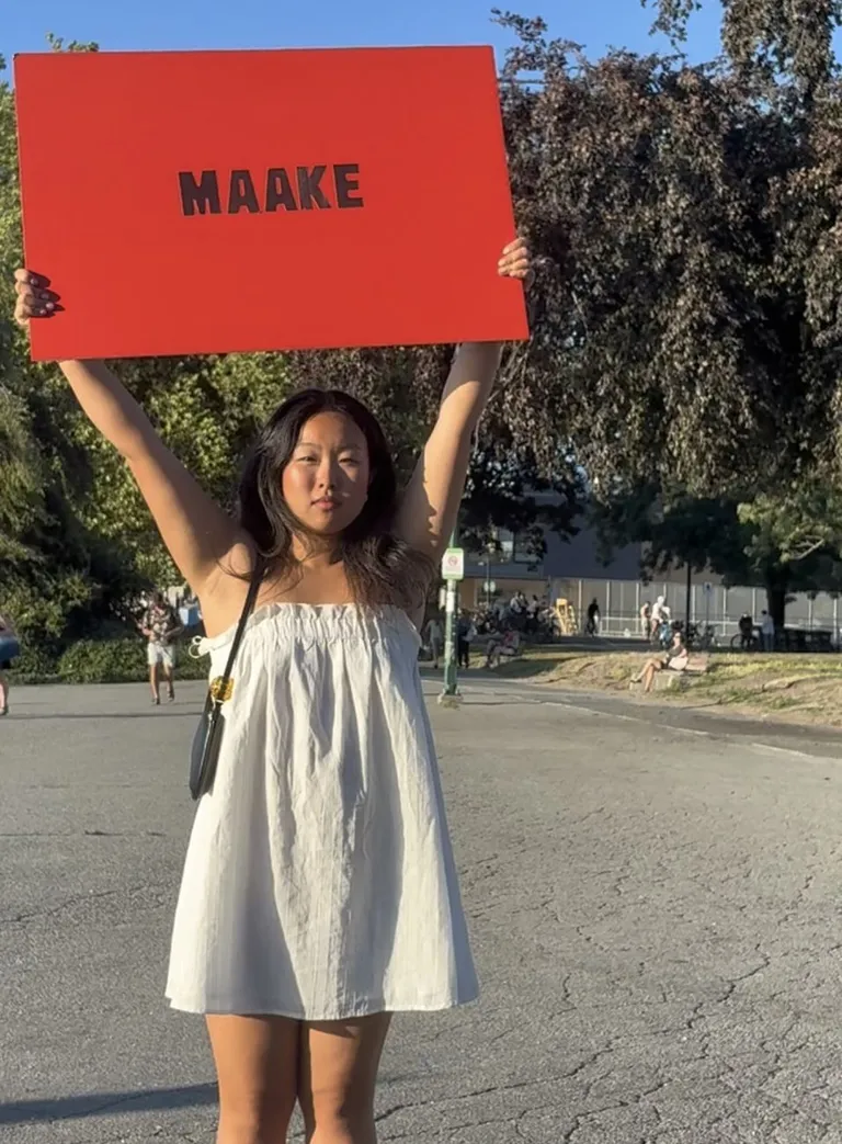 A woman in a white dress holding a red Maake sourdough crackers sign above her head at golden hour in a park