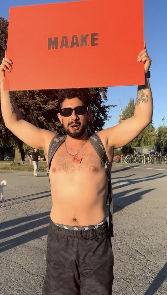 A grinning man in sunglasses holding a red Maake sign overhead at Kits Beach